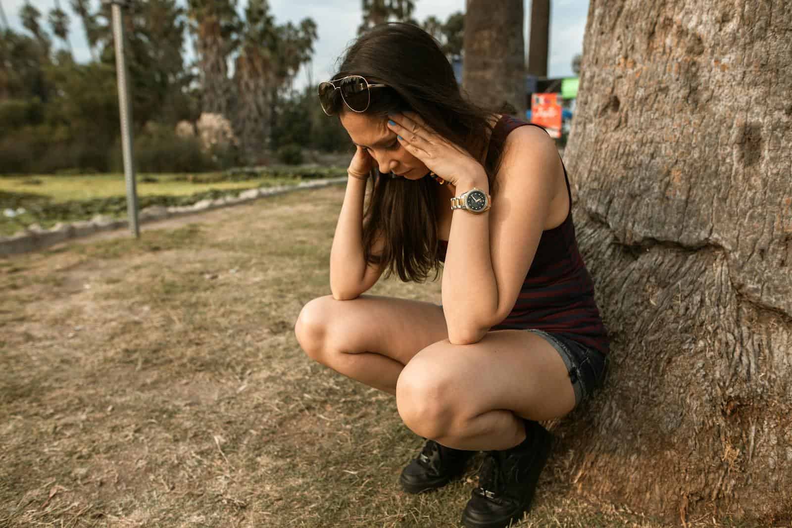 Indigestion, guts, bloating, gas, acid reflux, health, functional medicine, wellness, mind gut connection, A woman kneels by a tree holding her head, expressing stress or anxiety in an outdoor park.